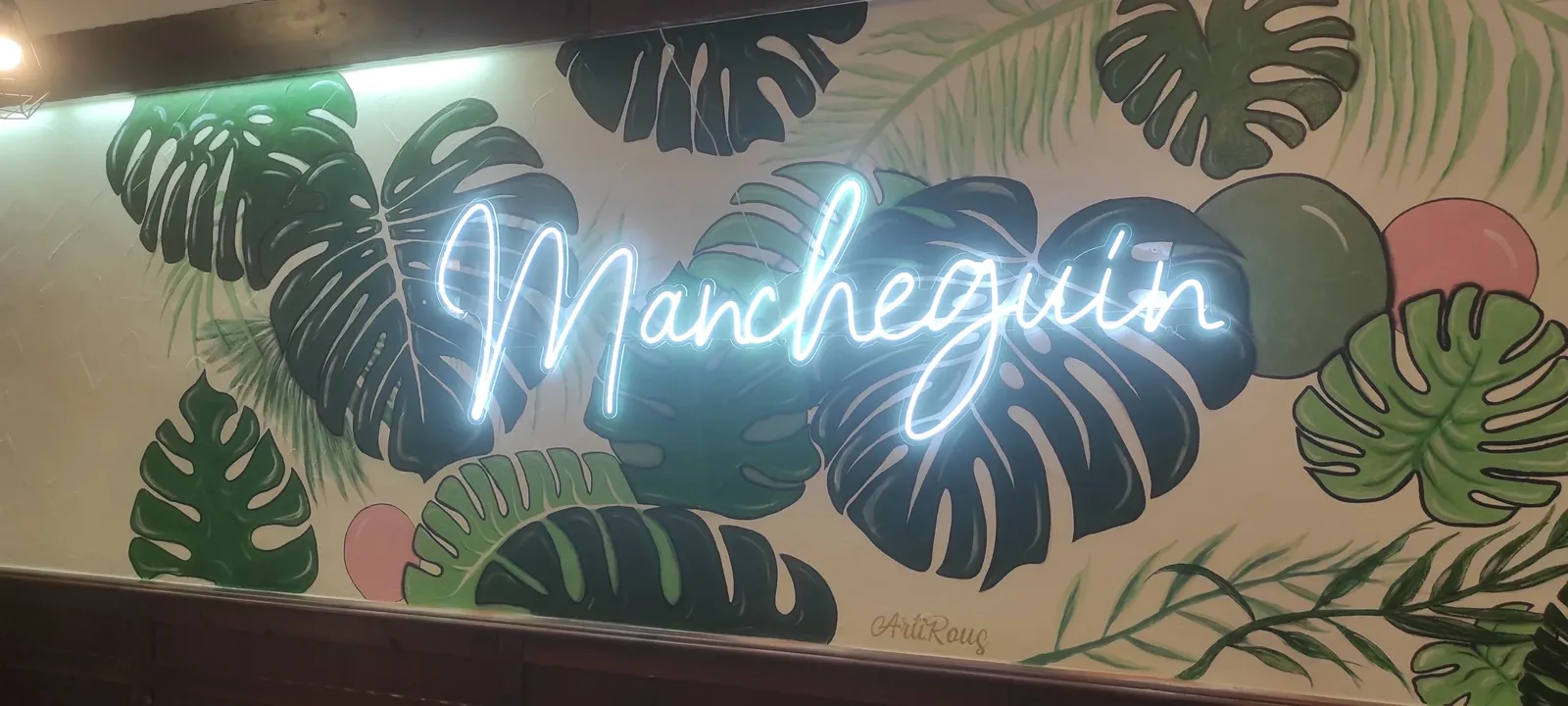 Neón LED con el nombre Mancheguín sobre una pared pintada con hojas tropicales